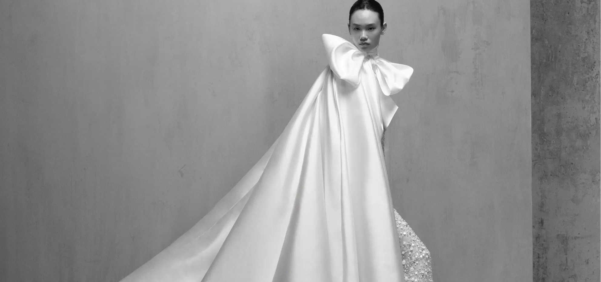 Woman in a glamorous gown with a large bow standing on steps against a plain wall.